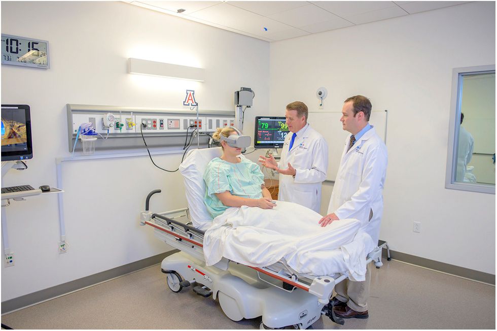 Two male doctors standing over a woman, in a hospital gown, in a hospital bed, wearing VR goggles