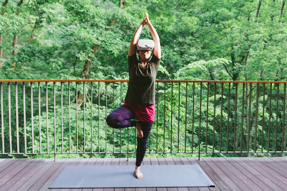 A woman in a yoga pose, with her hands above her head on a mat on a porch, with green trees behind her