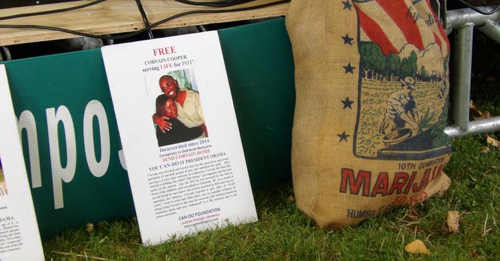 A sign propped between a lawn and a stage with the words "Free Corvain Cooper serving LIFE for POT!" next to a burlap bag with the word marijuana on it
