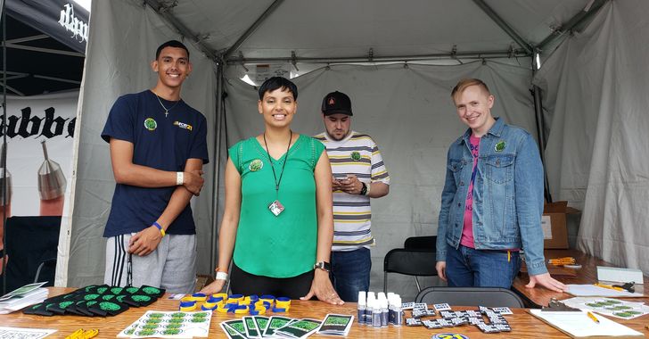 Image: Four people standing behind a table full of assorted objects in front of a white tent