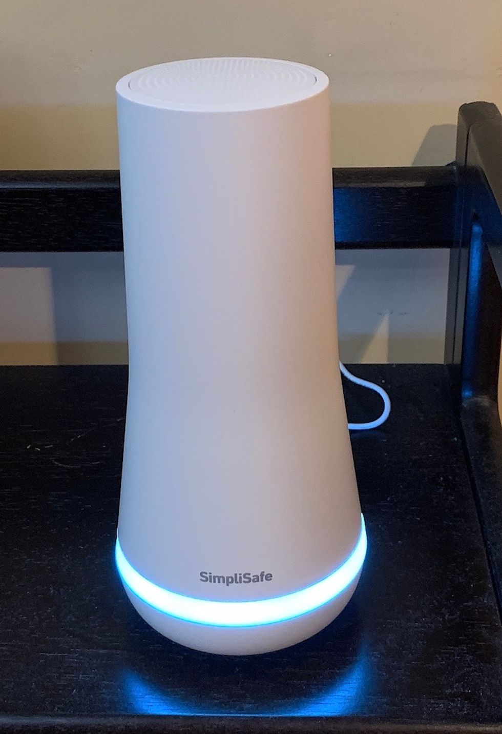 A tall white plastic device, with a blue light ring at the base and the word "SimpliSafe" written across the bottom