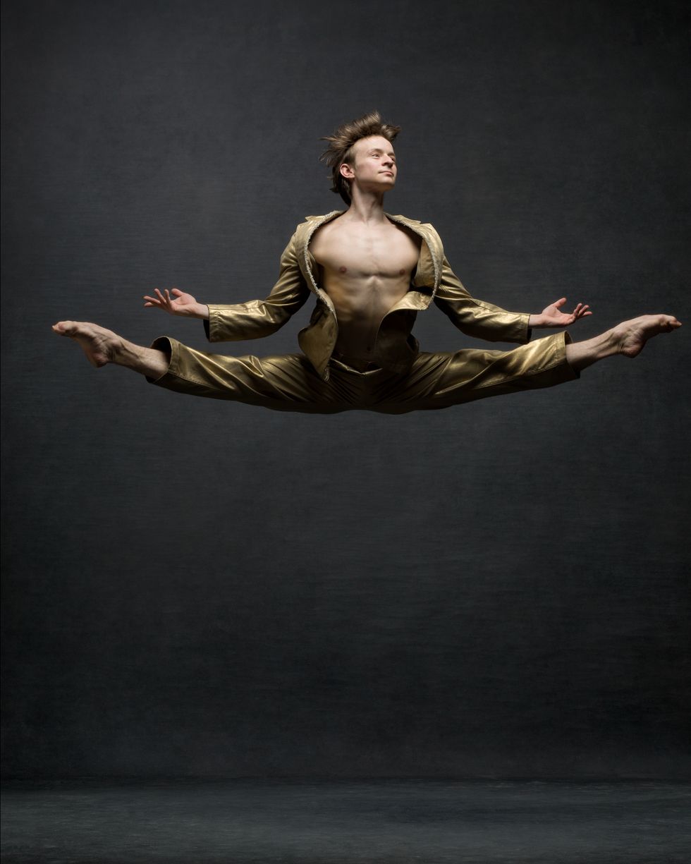 A male dancer is jumping high off the ground with split legs. He wears a gold vintage suit.