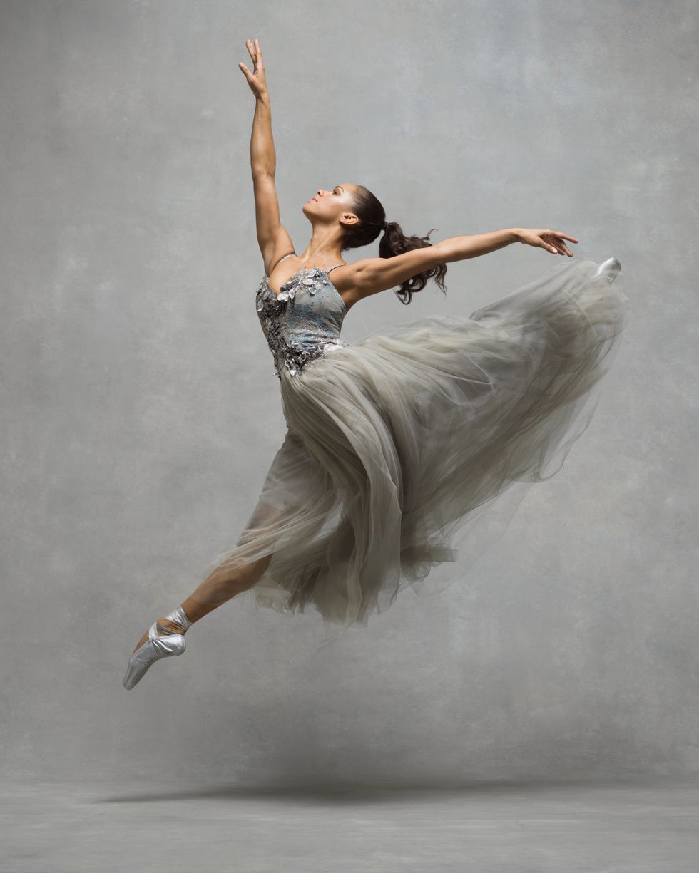 A female dancer jumps of the ground in profile, splitting her legs and looking towards a high front arm. She wears a long silver and gray gown and silver pointe shoes.