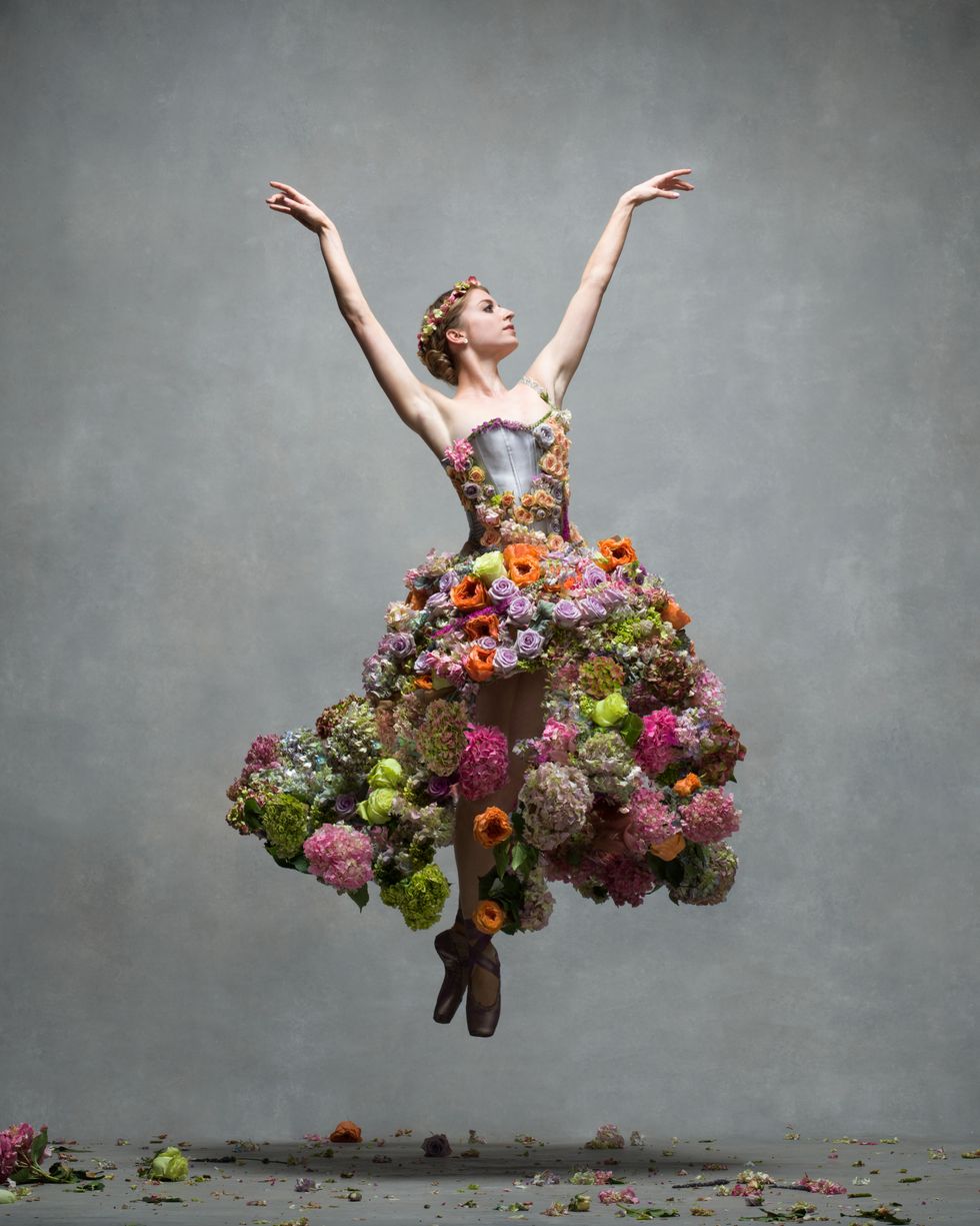 A female dancer jumps off the ground with two pointed feet and arms high over her head. Her dress has a colorful skirt made from real flowers, and there are flowers strewn on the ground.
