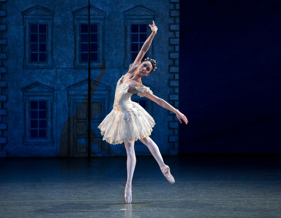 A female dancer stands onstage, balancing on one leg with her other leg raised behind her. She is wearing a white tutu and crown.