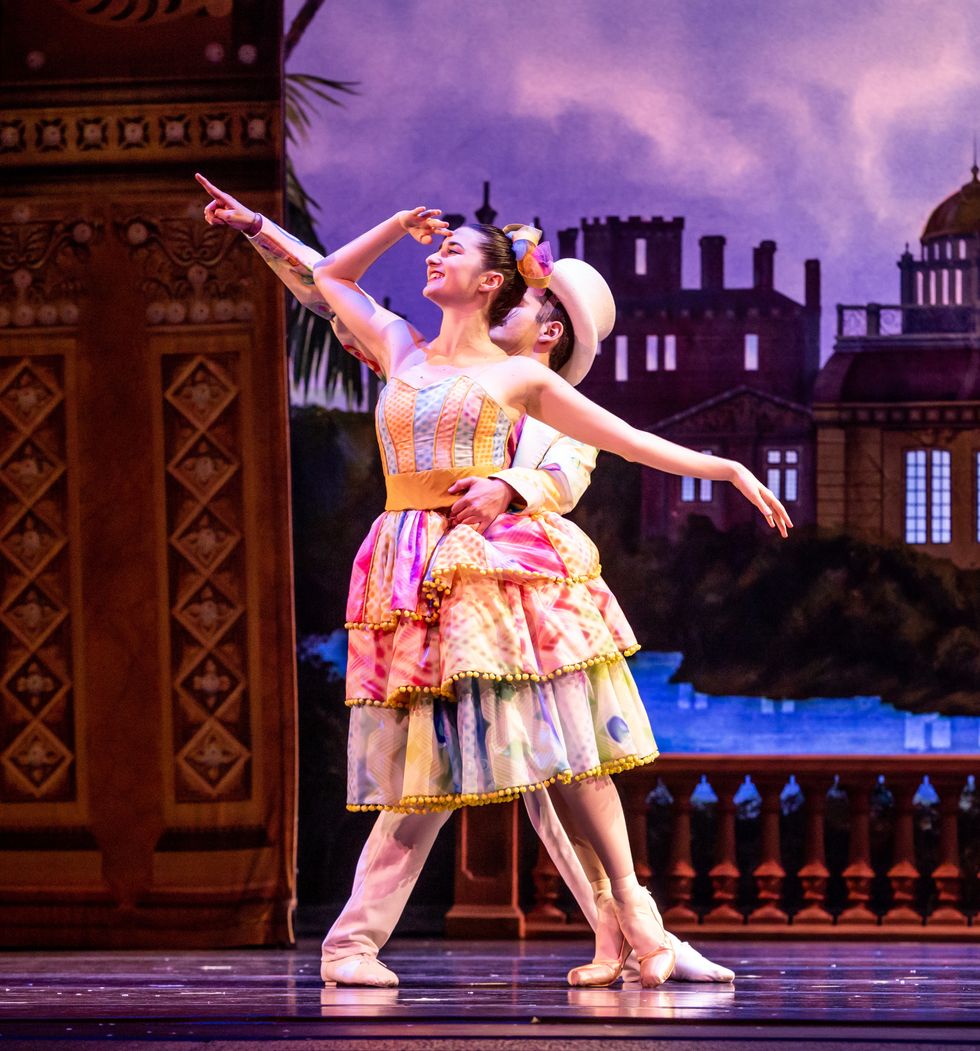 A male and female dancer pose close together. The woman is risingon pointe and wearing a pink, purple, yellow knee-length tutu. The man wears a white jacket and pants and has a top hat on.