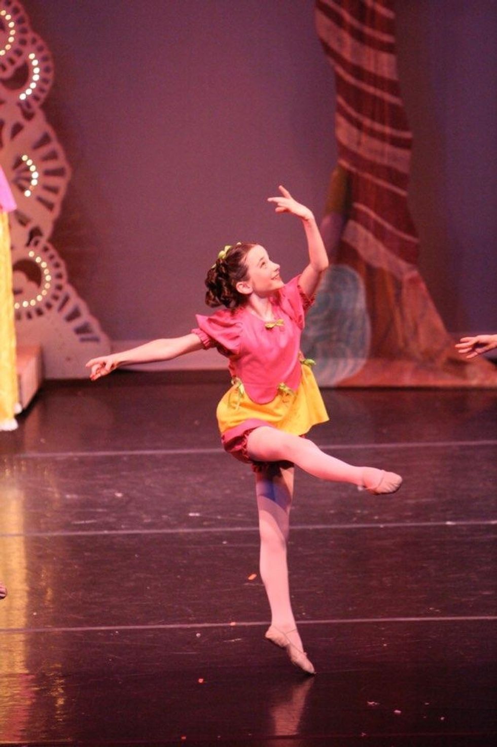 A young girl jumps lightly off one leg during an onstage performance, wearing a bright pink and yellow costume and tights.