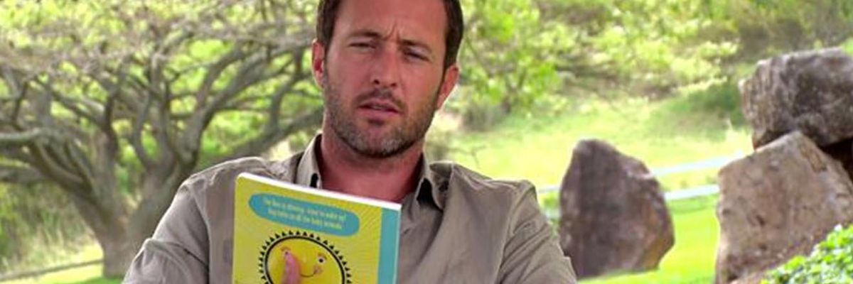 Hawaii Five-0's Alex O'Loughlin squints as he reads a childrens book in a park.