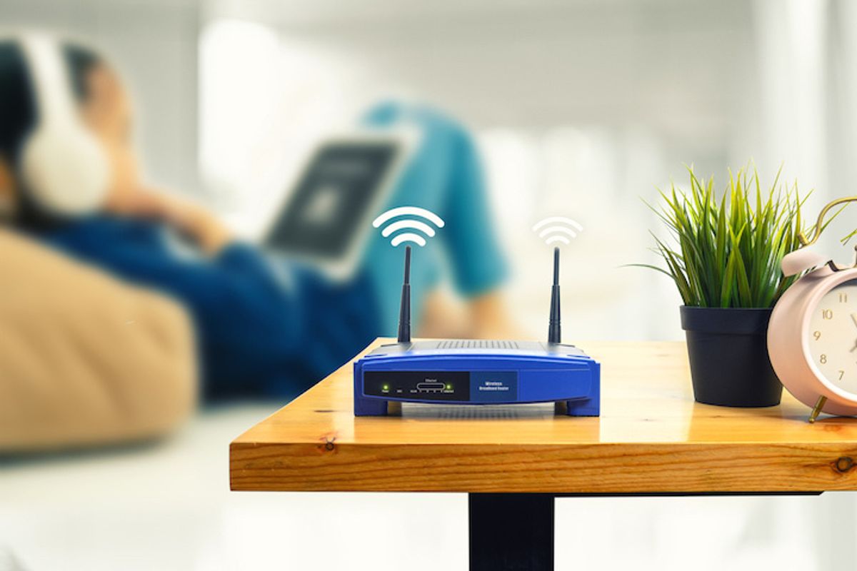 A router on a table with someone in the background on a tablet, wearing headphones