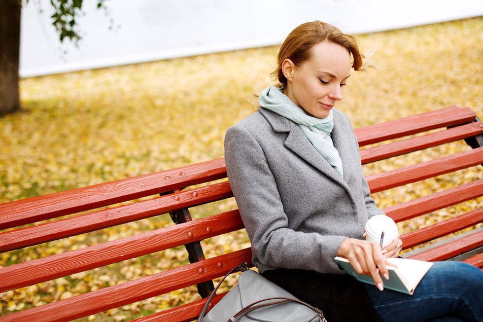 Working mom taking some time to herself, sitting on a bench in the park, drinking coffee and writing in a journal.