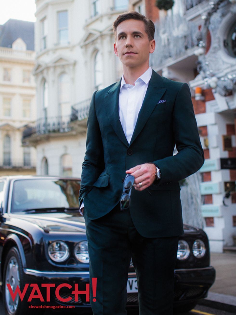 Brian Dietzen holding a pair of sunglasses and wearing a suit while standing in front of a Bentley