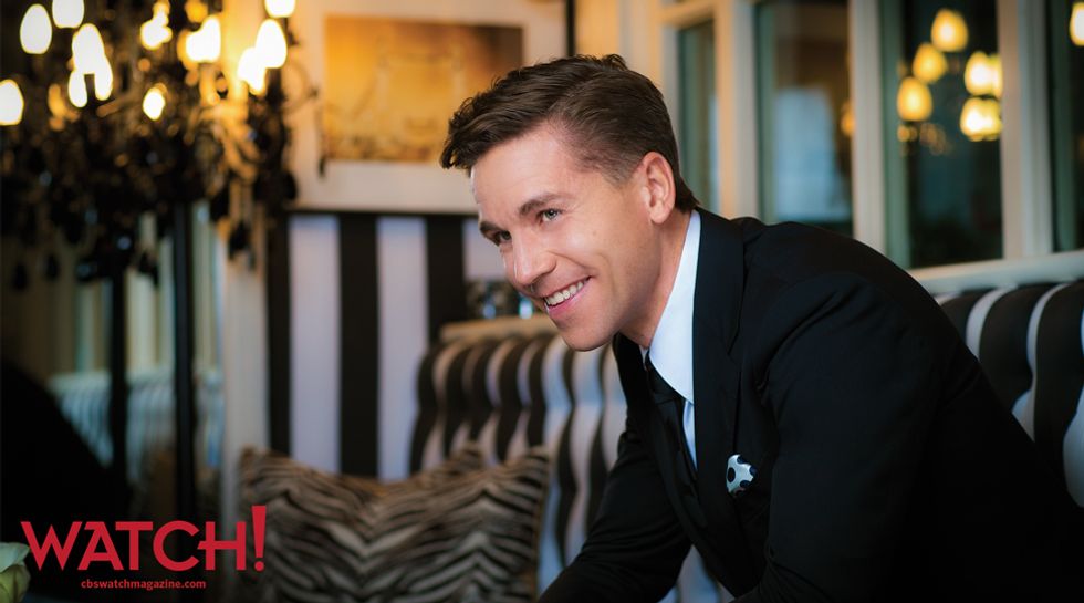 Brian Dietzen smiling off into the distance while sitting on a leopard print couch