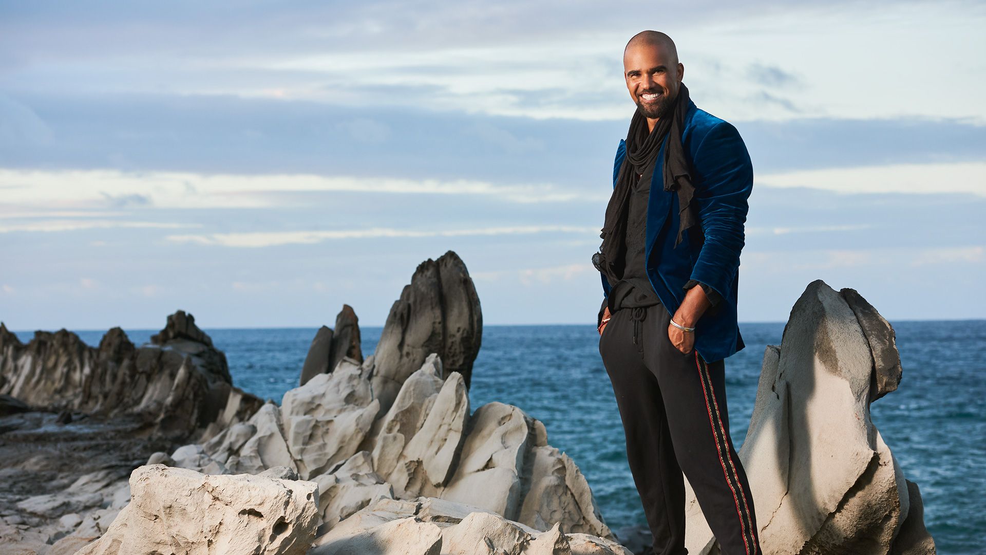 Shemar Moore in a blue velvet jacket standing on rocky terrain in front of the ocean.