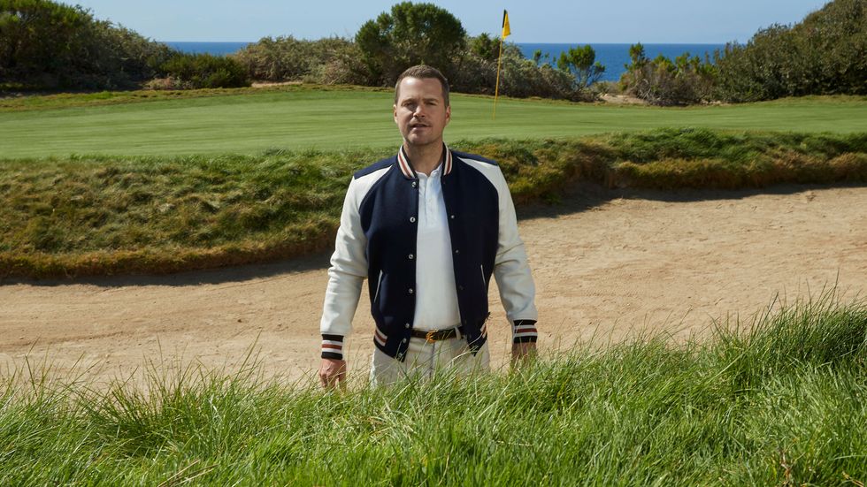 Chris O'Donnell wears a letterman's jacket as he stands in a sand trap of a golf course.