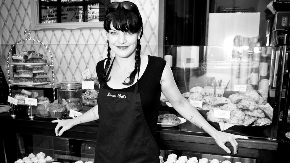 Black and white image of Actress Pauley Perrete wearing an apron in front of baked goods.