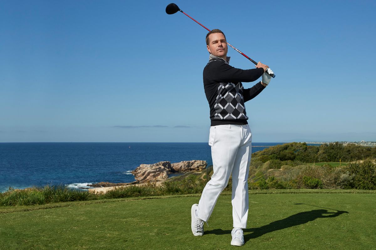 Chris O'Donnell swings a golf club while wearing a patterned golf sweater and white pants.