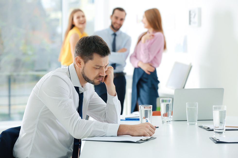 Young man looking sad at his desk as his co-workers laugh and talk behind him.