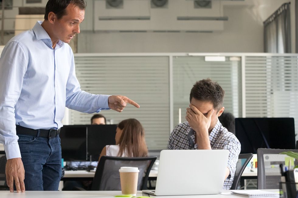 Young professional upset at his desk while his co-worker yells at him.