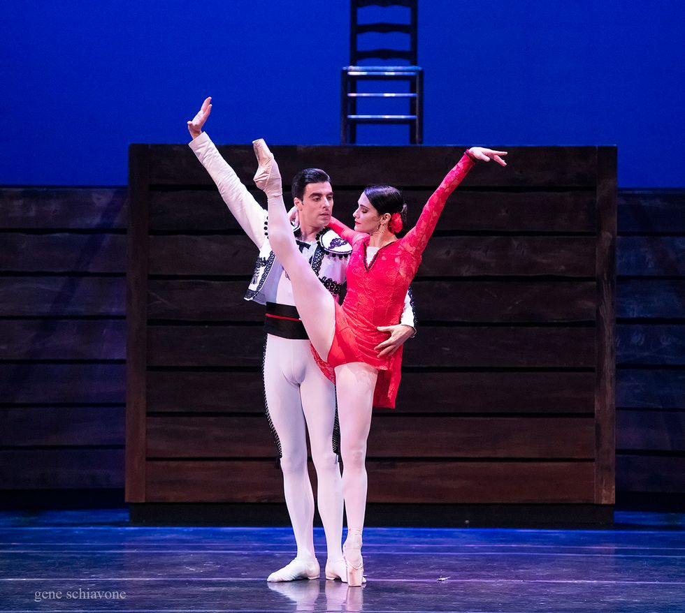 Sarah Lane, wearing a short red, lacy costume, kicks her right leg high in the air. Luis Ribagorda, wearing white tights and a white bolero jacket, holds on to her waist.