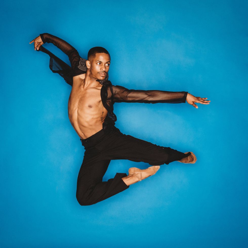 Roderick Phifer, dressed in black pants, ballet shoes and an open black, sheer shirt jumps in the air against a blue background.