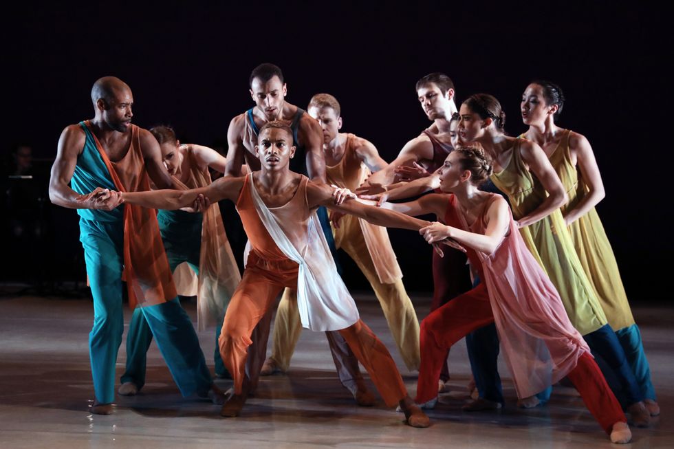 A group of dancers in brightly colored pants and tunics hold back Roderick Phifer, who stands in a parallel lunch with his arms spread in both directions.