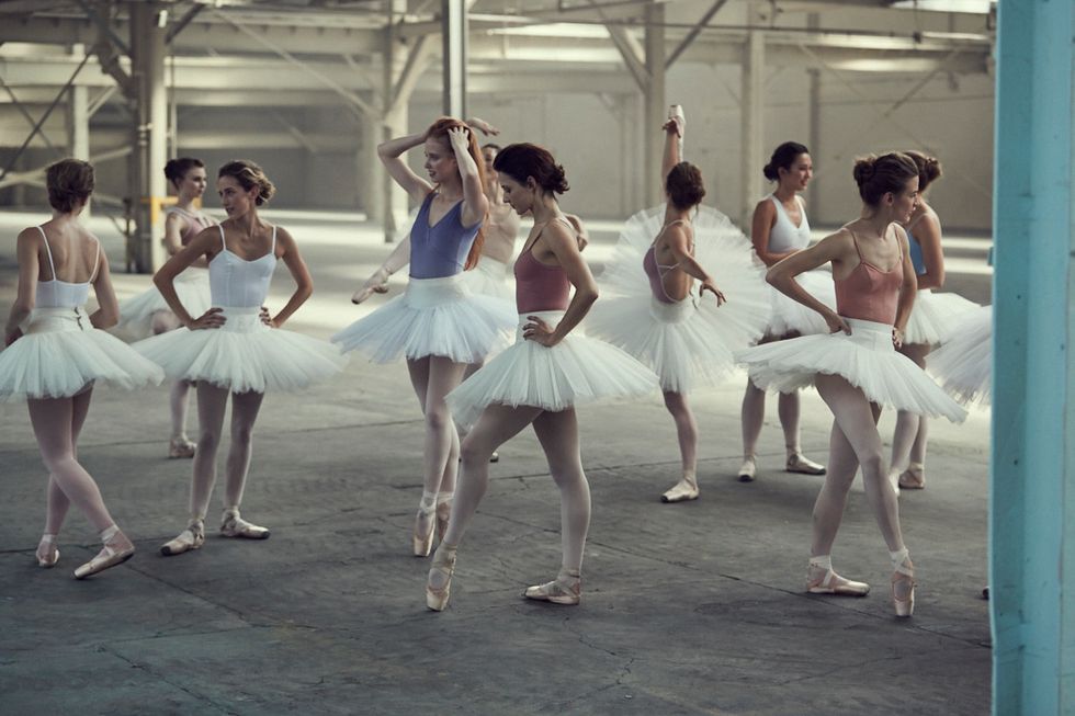 A number of ballerinas stand around a warehouse space wearing different colored leotards, white tutus, pink tights and pointe shoes.