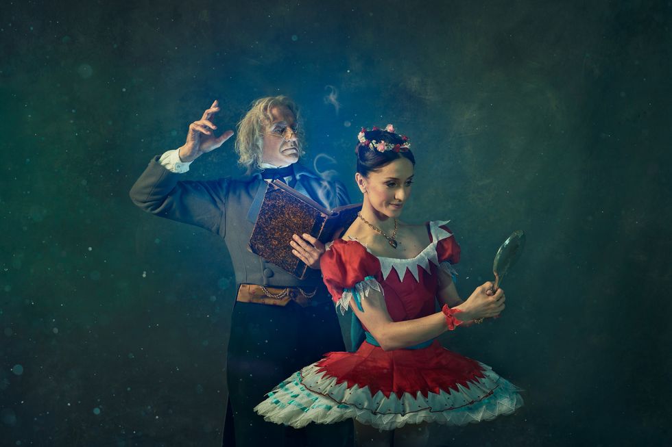 Marianela Nu\u00f1ez, as Swanilda, looks at her reflection in a handheld mirror, She wears a red tutu. Behind her, Gary Avis, as Dr. Coppelius, reads a book. He wears a gray jacket and white-haired wig.