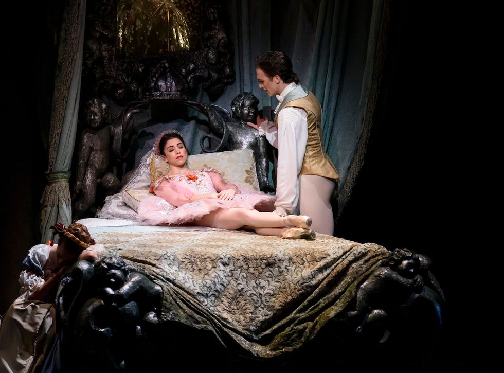 Yasmine Nagdhi, in a pink Sleeping Beauty tutu, lies on a bed while Matthew Ball, in a taupe silk jacket and tights, stands over her.