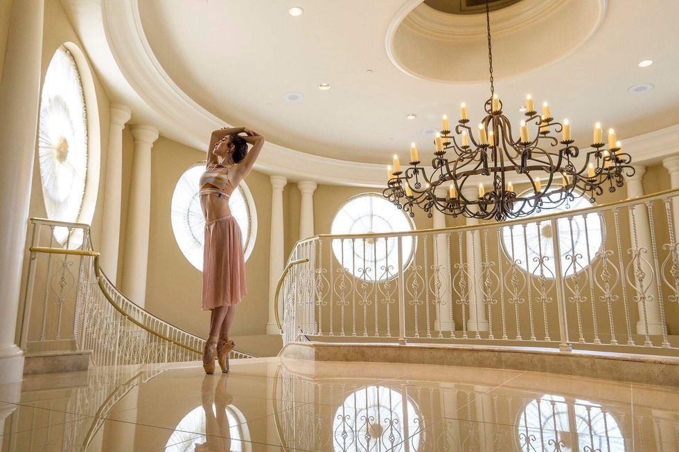 Isabell Mendez in a pink skirt, crop top and pointe shoes stands in an elaborate, fancy hall above a wrought iron staircase, with a large chandelier.