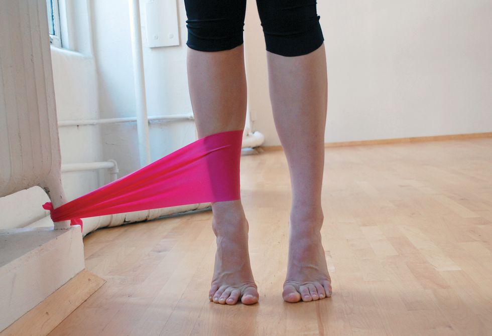 A dancer stands in black leggings on a wooden studio floor. We only see her legs. She is standing in relev\u00e9, and a pink elastic band attaches her ankle to the base of a white radiator.