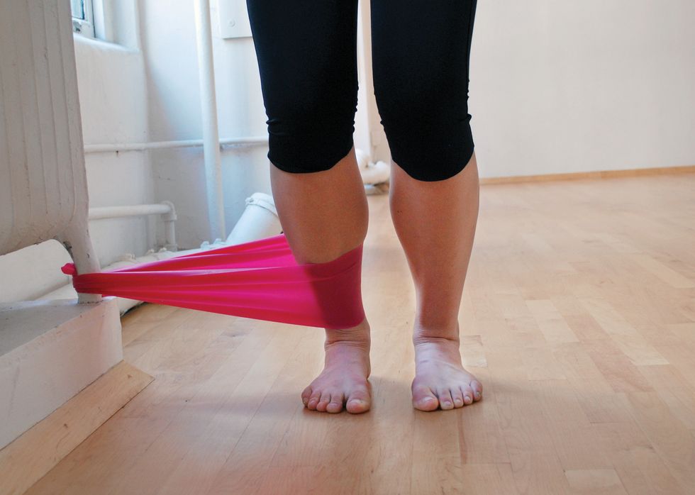 A dancer stands in black leggings on a wooden studio floor. We only see her legs. She is standing in pli\u00e9, and a pink elastic band attaches her ankle to the base of a white radiator.