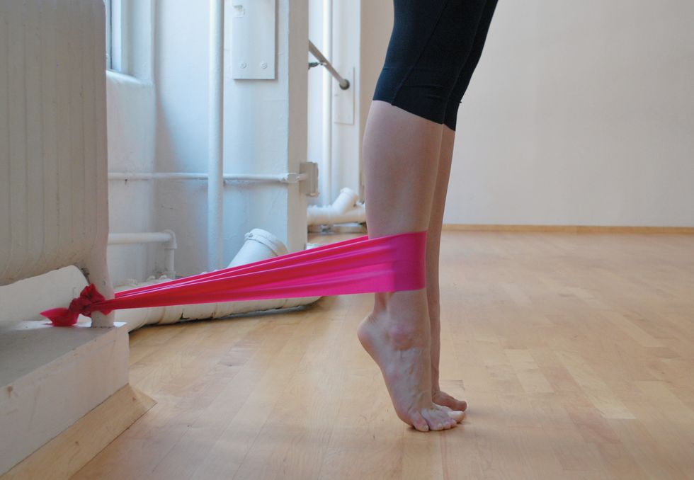 A dancer stands in black leggings on a wooden studio floor. We only see her legs. She is standing in relev\u00e9, and a pink elastic band attaches her ankle to the base of a white radiator. We see her legs in profile..