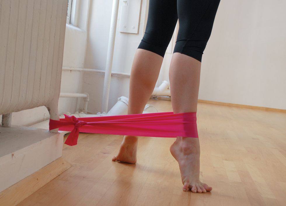 A dancer stands in black leggings on a wooden studio floor. We only see her legs. She is standing in relev\u00e9 turned out, and a pink elastic band attaches her ankle to the base of a white radiator.