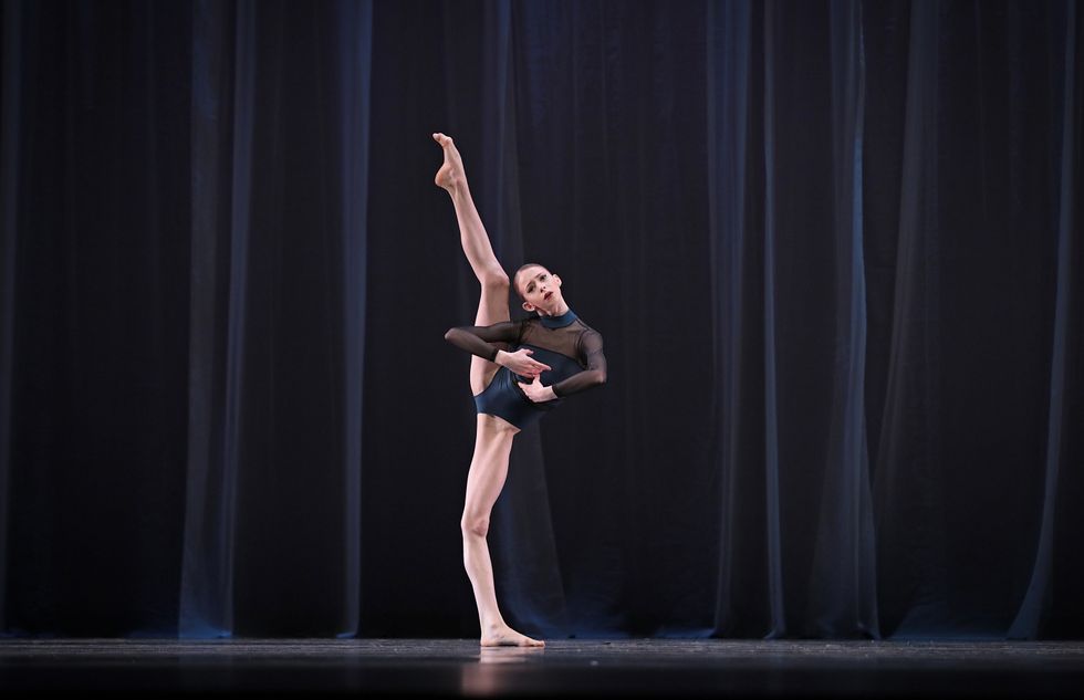 A dancer stands in a navy and black sheer leotard with one leg extended way above her head.