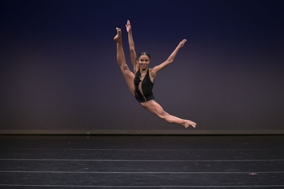 Madison Brown, in a black leotard costume, leaps in the air with her legs over extended.