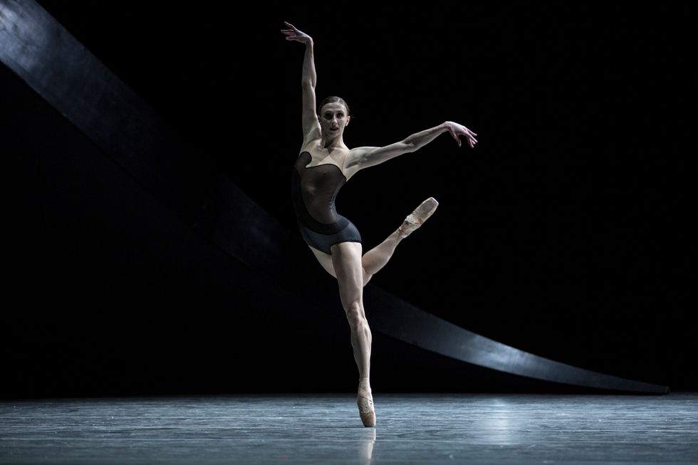 Macy onstage in a dark background wearing a black leotard costume and pointe shoes. One leg is extended in a kind of attitude behind her.