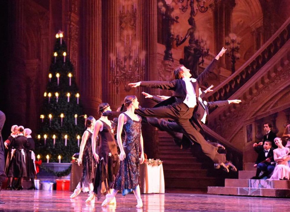 Chandler Proctor, in a black tuxedo costume, splits his legs into sissonne during the Nutcracker's party scene. He is surrounded by other dancers in black dresses and tuxedos.