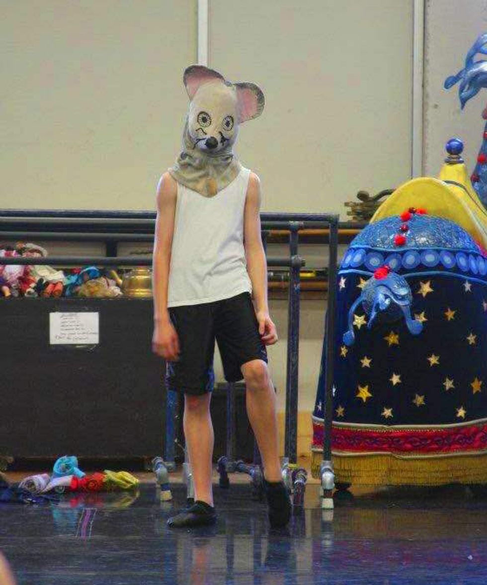 Sam Ainley, in a mouse headpiece, white sleeveless T-shirt, black shorts and black ballet shoes, listens to rehearsal notes in a cluttered dance studio.