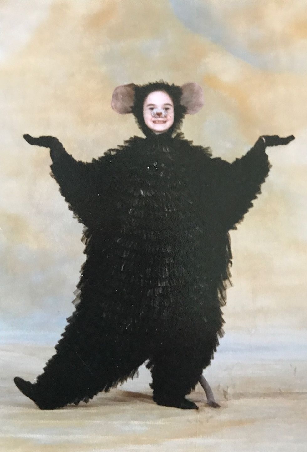 Elizabeth Powell, as a child, poses in a fluffy black mouse costume and headpiece. She wears makeup of a mouse nose and whiskers on her face.