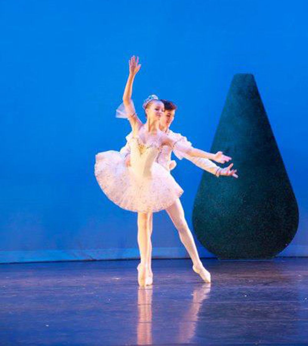 A young Charles Proctor, wearing a white jacket and tights, stands behind a teenage student ballerina in a white tutu and holds her waist so she can balance on pointe.