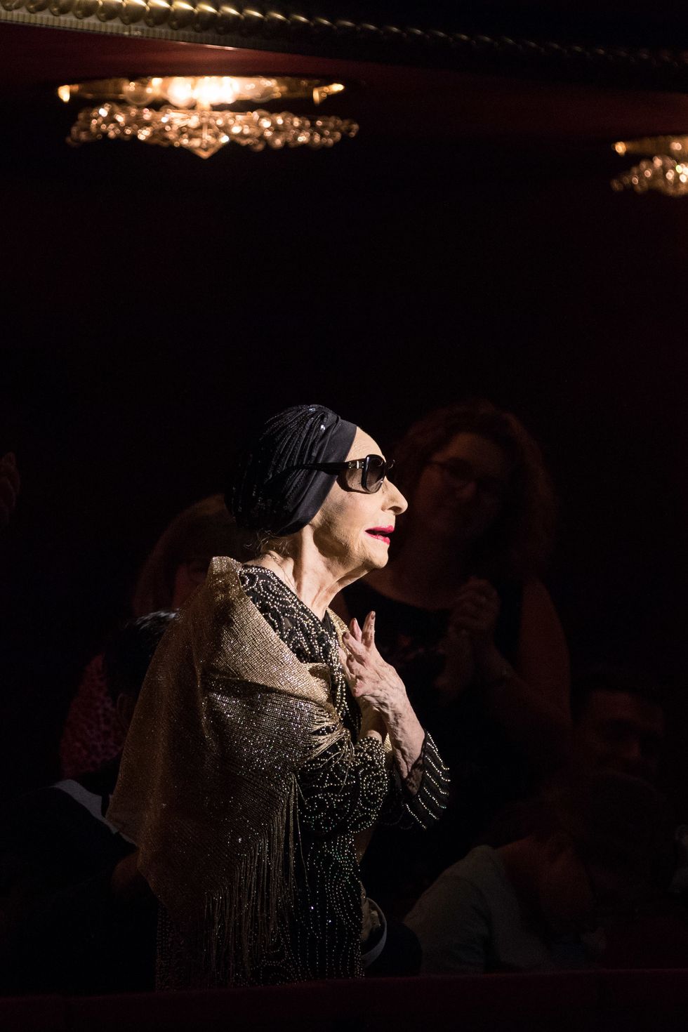 An elderly woman wearing sunglasses and a black headscarf stands in a spotlight, taking a bow.
