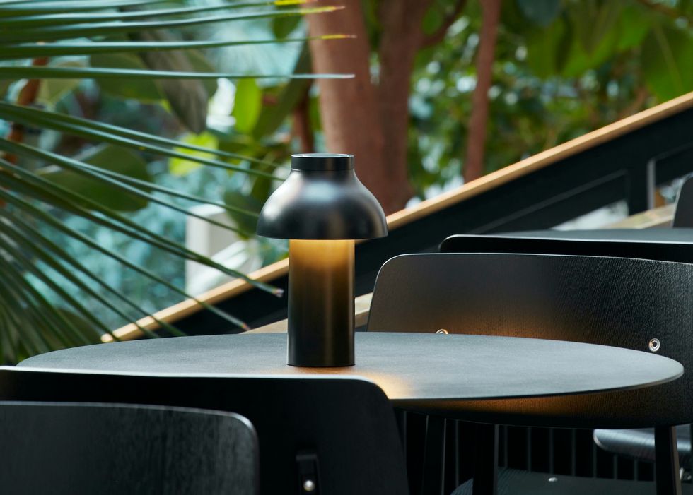 A brown and black lamp from Hay on an outdoor table surrounded by greenery