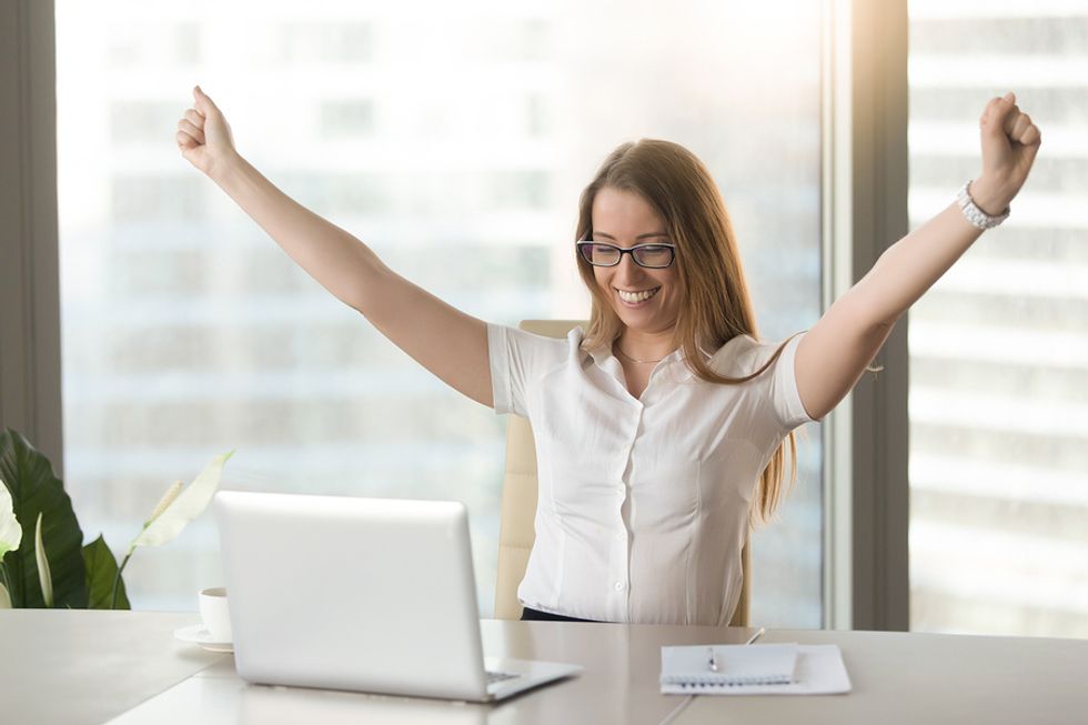 Young professional woman cheering in excitement at her desk because she's excited to be working a job she's passionate about.