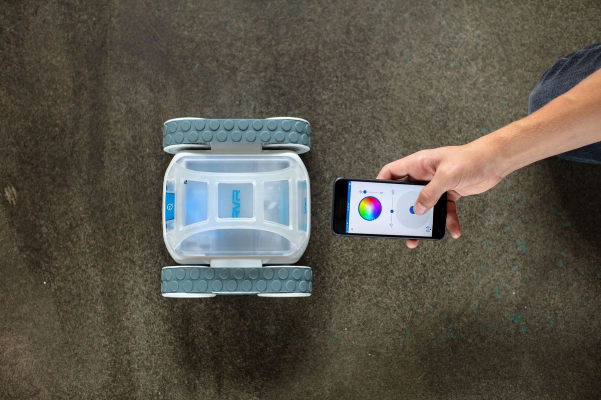 An RVR robot in white and clear plastic from Sphero next to a hand holding a smartphone