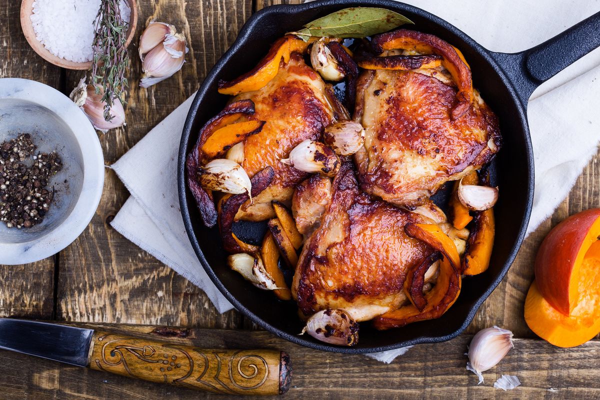 A cast iron skillet of chickens and garlic cloves on a wood table with spices and a knife