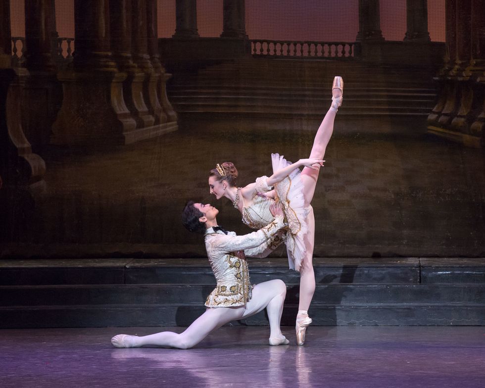 A male and female ballet dancer pose together onstage. The male , wearing a prince costume, is on his knee holding the waist of the ballerina, who wears a tutu and leans forward toward him on pointe.