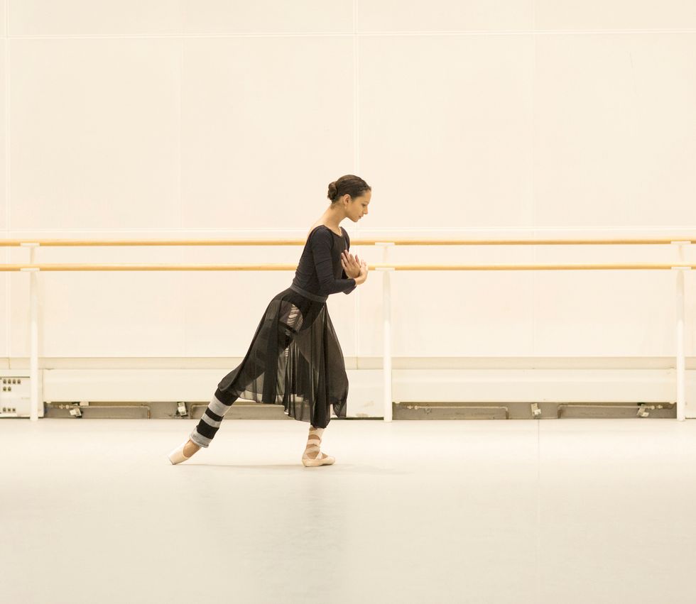 In a black leotard and long black skirt, pointe shoes and striped legwarmers, Francesca Haywards leans forward in a back tendu.