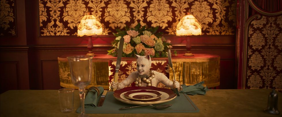 Francesca Hayward in a white cat costume sits at a giant, ornate table holding a huge fork and knife.