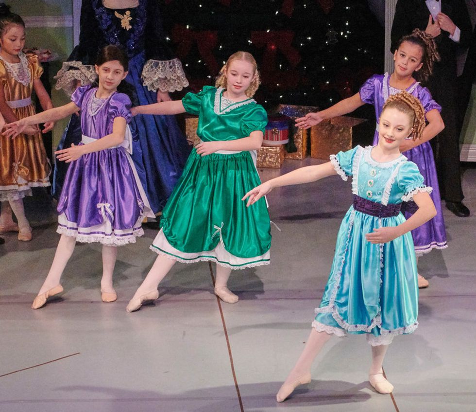 A stage photo of Nutcracker of four young girls dressed in colorful party dresses and soft shoes. Stella Jaworski wears a purple dress and a brown wig.