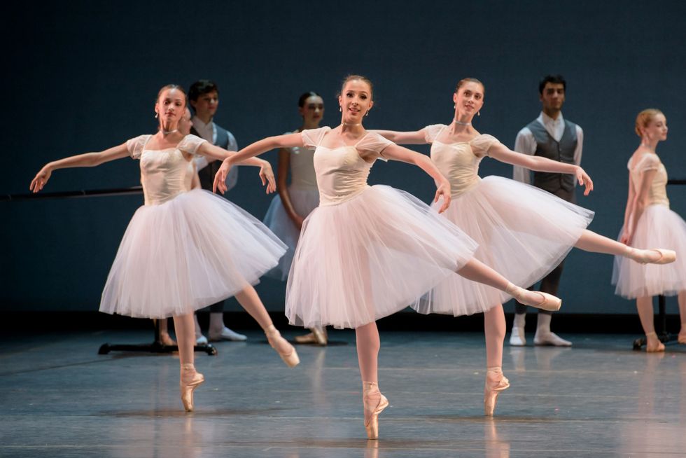 Three female Boston Ballet students pose in arabesque onstage, wearing long white tutus.